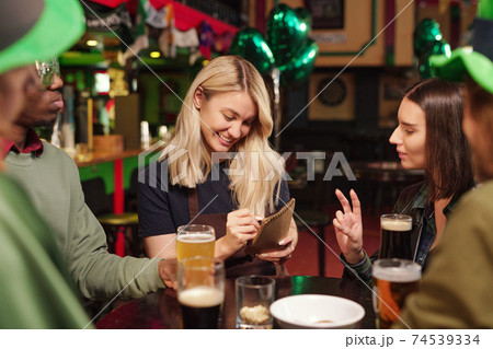 Happy young blond waitress in workwear writing down orders of clients in notepad Happy young blond waitress in workwear writing down orders of clients in notepad 74539334