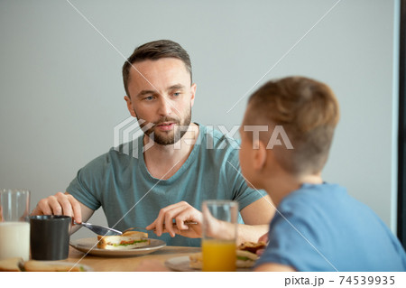 Serious young man eating homemade sandwich for breakfast and talking to son 74539935