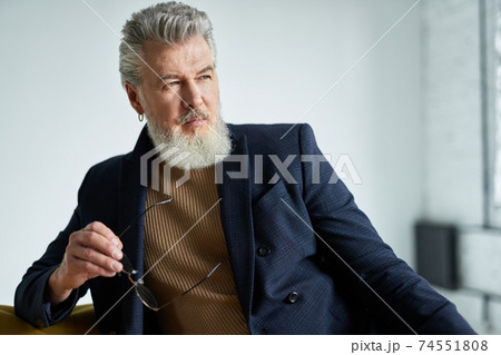 Portrait of stylish serious middle aged man with beard looking away, holding glasses while sitting on sofa indoors Portrait of stylish serious middle aged man with beard looking away, holding glasses while sitting on sofa indoors 74551808
