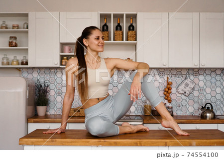 Athletic woman in sportswear posing on the table top of a light modern kitchen Athletic woman in sportswear posing on the table top of a light modern kitchen 74554100