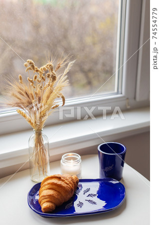Handmade blue clay plate with croissant on it. Vase with wheat and candles. Handmade blue clay plate with croissant on it. Vase with wheat and candles. 74554779