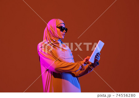 Beautiful arab woman posing in stylish hijab isolated on brown studio background in neon light. Fashion, beauty, style concept Beautiful arab woman posing in stylish hijab isolated on brown studio background in neon light. Fashion, beauty, style concept 74556280