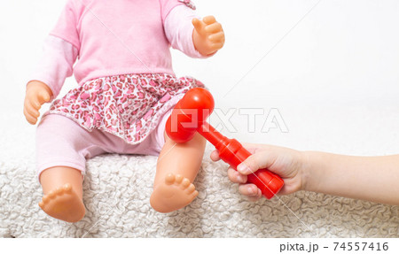 A little girl plays with a doll, and with a small neurological hammer, hits the knee of the doll. Child neurology concept, reflex check, copy space A little girl plays with a doll, and with a small neurological hammer, hits the knee of the doll. Child neurology concept, reflex check, copy space 74557416