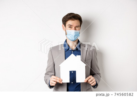 Real estate and covid-19 concept. Cheerful young man in medical mask and suit winking at camera, advertising new flats, showing paper house cutout Real estate and covid-19 concept. Cheerful young man in medical mask and suit winking at camera, advertising new flats, showing paper house cutout 74560582