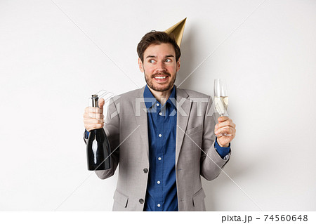 Celebration and holidays concept. Happy birthday guy in suit and party hat drinking champagne, holding bottle and glass, smiling and looking aside Celebration and holidays concept. Happy birthday guy in suit and party hat drinking champagne, holding bottle and glass, smiling and looking aside 74560648