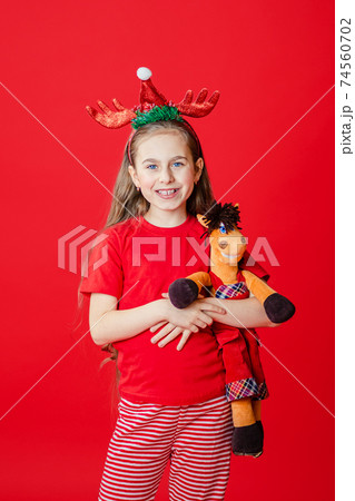 Portrait of a funny cheerful girl with a bandage of horns on her head hugging a teddy horse in Christmas pajamas isolated on a bright red background. Portrait of a funny cheerful girl with a bandage of horns on her head hugging a teddy horse in Christmas pajamas isolated on a bright red background. 74560702