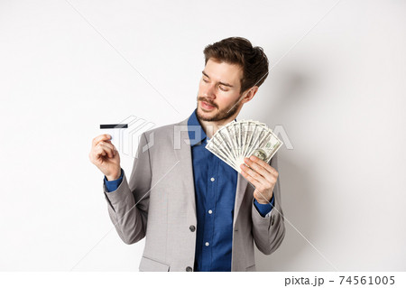 Successful man holding dollar bills and looking at plastic credit card, pleased with bank, standing on white background in business suit 74561005
