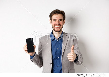 E-commerce and online shopping concept. Satisfied businessman in suit showing empty smartphone screen and thumb-up, praise good deal, white background E-commerce and online shopping concept. Satisfied businessman in suit showing empty smartphone screen and thumb-up, praise good deal, white background 74562780