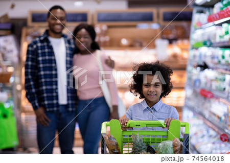 Happy little black girl with shopping trolley and her parents buying goods at modern supermarket, free space Happy little black girl with shopping trolley and her parents buying goods at modern supermarket, free space 74564018