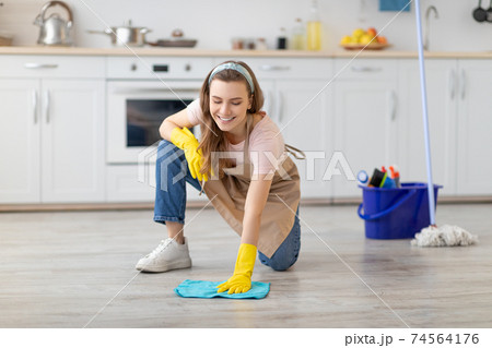 Professional cleaning service. Cheerful young lady in apron and rubber gloves washing floor in modern kitchen Professional cleaning service. Cheerful young lady in apron and rubber gloves washing floor in modern kitchen 74564176