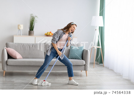 Pretty millennial woman singing while wiping floor at home, using mop as microphone, having fun during domestic duties Pretty millennial woman singing while wiping floor at home, using mop as microphone, having fun during domestic duties 74564338