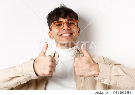 Close-up of satisfied happy guy showing thumbs up and smiling with white teeth, wearing eyewear, standing on white background 74566198
