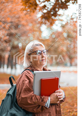 Caucasian blonde woman holding notebook folders in her hands walking in the park. Teacher in casual clothes, glasses and with a backpack, Education, studying foreign languages concept 74567298