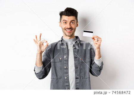 Very good. Smiling guy with plastic credit card showing okay sign, smiling satisfied, recommend bank, standing on white background 74568472