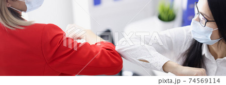 Two businesswomen in protective face masks touching elbows in office 74569114