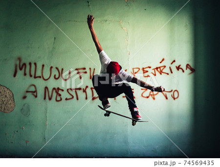 Young skateboarder caught in peak moment of his stunt with arms outstretched, board lightly touching shoes. In background wall of room in abandoned house with sprayed text. Natural light conditions. 74569435