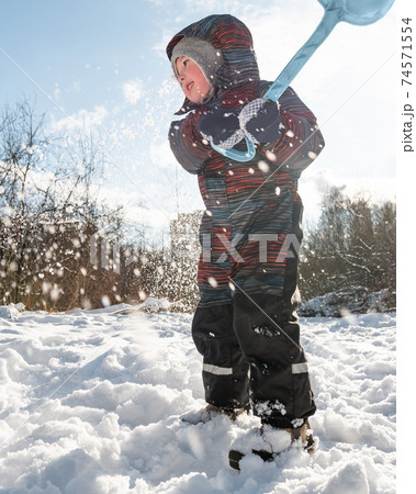Kid makes snowfall with his toy shovel. Child plays with snow. Leisure activity outdoor at winter season. Happy boy in warm jumpsuit. 74571554