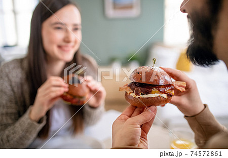 Unrecognizable young couple in love eating hamburgers indoors at home. Unrecognizable young couple in love eating hamburgers indoors at home. 74572861