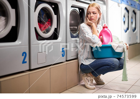 Laundry interior, young caucasian woman sitting near washing machines and looking up sadly Laundry interior, young caucasian woman sitting near washing machines and looking up sadly 74573599