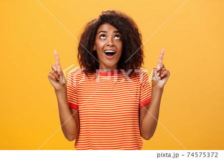 Optimistic happy african american girl saying thanks to god looking and pointing up pleased and carefree starting day in productive way being delighted with lucky chance over orange background Optimistic happy african american girl saying thanks to god looking and pointing up pleased and carefree starting day in productive way being delighted with lucky chance over orange background 74575372