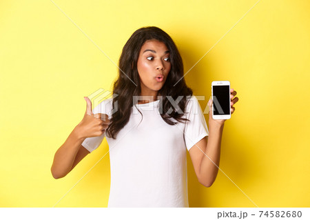 Portrait of good-looking african american female model, recommending an app, showing mobile phone and look excited, standing over yellow background Portrait of good-looking african american female model, recommending an app, showing mobile phone and look excited, standing over yellow background 74582680
