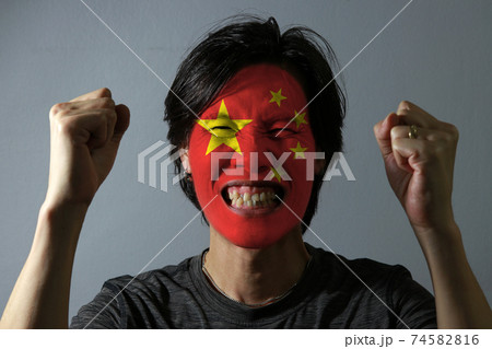 Cheerful portrait of a man with the flag of the China painted on his face on grey background. Cheerful portrait of a man with the flag of the China painted on his face on grey background. 74582816