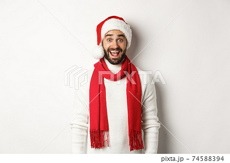 Christmas holidays. Bearded man looking surprised at camera, wearing santa party hat and red scarf, standing against white background Christmas holidays. Bearded man looking surprised at camera, wearing santa party hat and red scarf, standing against white background 74588394