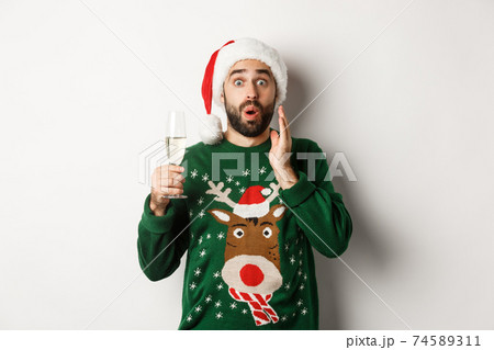 Christmas party and holidays concept. Surprised man in Santa hat celebrating New Year, drinking champagne and rejoicing, standing over white background 74589311
