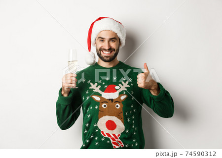 Christmas party and holidays concept. Satisfied guy in Santa hat and sweater showing thumbs up and drinking glass of champagne, standing over white background Christmas party and holidays concept. Satisfied guy in Santa hat and sweater showing thumbs up and drinking glass of champagne, standing over white background 74590312
