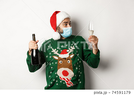 Concept of covid-19 and Christmas holidays. Man in medical mask and Santa hat enjoying glass of champagne, celebrating New Year, standing over white background 74591179