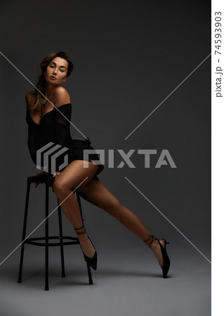 Young attractive brunette sexy woman sits on chair in erotic black cocktail dress. Gray background Young attractive brunette sexy woman sits on chair in erotic black cocktail dress. Gray background 74593903