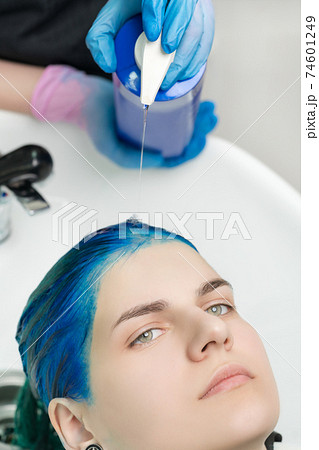 Close-up view of hairdresser squeezes shampoo and conditioner from tube into head of woman with blue hair while washing hair in special sink Close-up view of hairdresser squeezes shampoo and conditioner from tube into head of woman with blue hair while washing hair in special sink 74601249