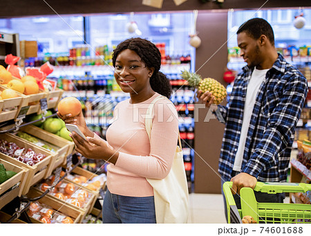 Happy black lady checking shopping list on mobile phone while choosing fruits with her boyfriend at supermarket Happy black lady checking shopping list on mobile phone while choosing fruits with her boyfriend at supermarket 74601688