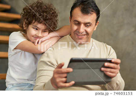 Middle aged latin father looking cheerful, holding tablet pc while spending time together with his son, sitting on the stairs at home 74603153