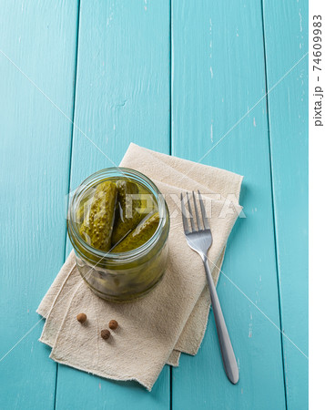 Crunchy pickled cornichons in a glass jar and fork over blue wooden table.  74609983