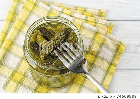 Delicious pickled cornichon on a fork over full glass jar against checkered napkin on a table. Delicious pickled cornichon on a fork over full glass jar against checkered napkin on a table. 74609988