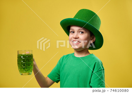 Smiling boy in green leprechaun Irish hat holds a glass with green drink and poses to the camera. Saint Patrick's Day. Isolated on yellow background. 74610080