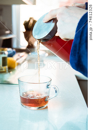Close up man in home bathrobe pouring hot tea from glass teapot into cup on the kitchen table. Process brewing tea in the morning. A cup of freshly brewed black tea, warm soft light. Selective focus. 74612326