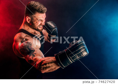 Bearded tattooed sportsman muay thai boxer in black undershirt and boxing gloves fighting on dark background with smoke. 74627420