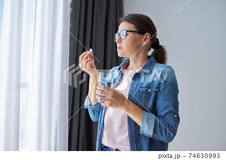 Mature middle-aged woman at home holding pill and glass of fresh water Mature middle-aged woman at home holding pill and glass of fresh water 74630993