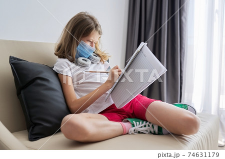 Child girl in face mask sitting on sofa with school notebook Child girl in face mask sitting on sofa with school notebook 74631179