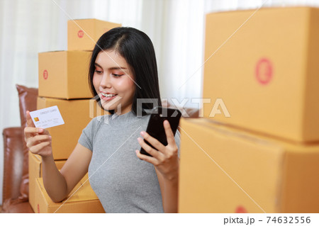 Young asian smart and active business woman in casual dress holding credit card and working from home with smartphone and online purchase shopping order box packaging (new normal concept) 74632556