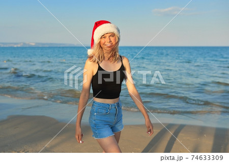 Beautiful girl in Santa hat walking along beach. Christmas and New Year holidays Beautiful girl in Santa hat walking along beach. Christmas and New Year holidays 74633309