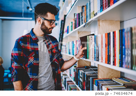 Successful joyful man student holding positive book while standing near shelves in university library 74634674