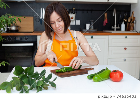 Young beautiful woman in apron sitting at table, cooking at home, chopping vegetables by knife 74635403