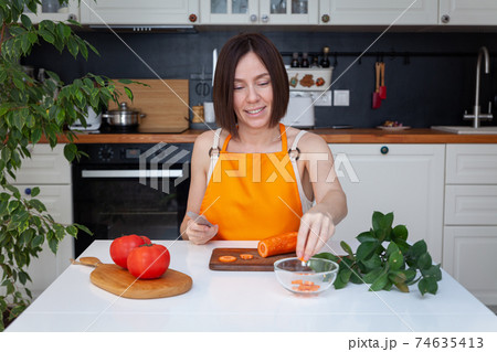 Young beautiful woman in apron sitting at table, cooking at home, chopping vegetables by knife Young beautiful woman in apron sitting at table, cooking at home, chopping vegetables by knife 74635413