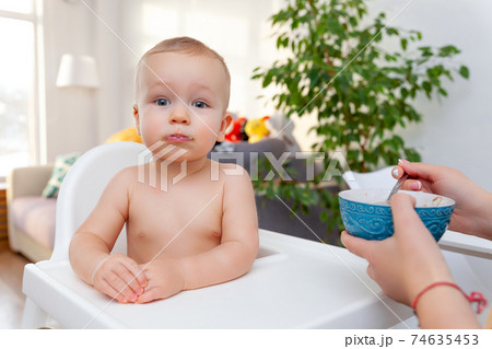 Mother's hand feeds funny cute happy blond baby toddler with spoon 74635453