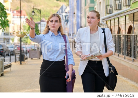 Two young business women walking and talking outdoors, city background 74635618