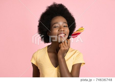 Pensive satisfied young african girl with afro hair, wearing yellow t-shirt, holding a lollipop, posing isolated on pink background. People, emotions, lifestyle concept. copy space. Sweets promo 74636748
