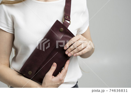 Girl in a white blouse with a leather red handmade bag over her shoulder. Designer dark red banana bag. Woman in a studio. Comfortable small bag for walking. 74636831